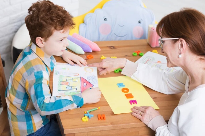 A child participates in an art therapy session with a therapist in Monmouth County, New Jersey. The therapist and child sit at a wooden table creating drawings and arranging colorful letters, using creative expression to support emotional development and communication in a safe, supportive setting.