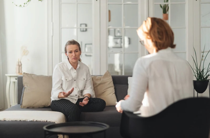 A woman speaks with a therapist during an individual therapy session at a counseling office in Monmouth County, New Jersey. The setting is calm and bright, with natural light, soft neutral tones, and a comfortable sofa that reflects a supportive, safe environment for mental health counseling.