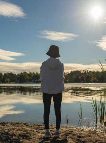 A person standing at the edge of a calm lake in Monmouth County, NJ, looking out over the water on a sunny day, wearing a hoodie and bucket hat with trees and homes in the distance.