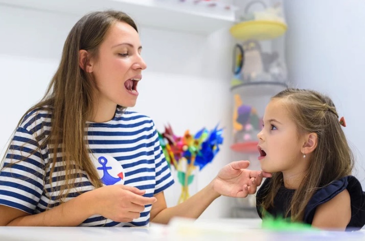 A speech therapist and a young girl sit across from each other in a bright therapy room, practicing mouth movements during a speech session. The therapist gently guides the child while colorful toys sit on shelves in the background, similar to offices used by local providers in Monmouth and Ocean County.