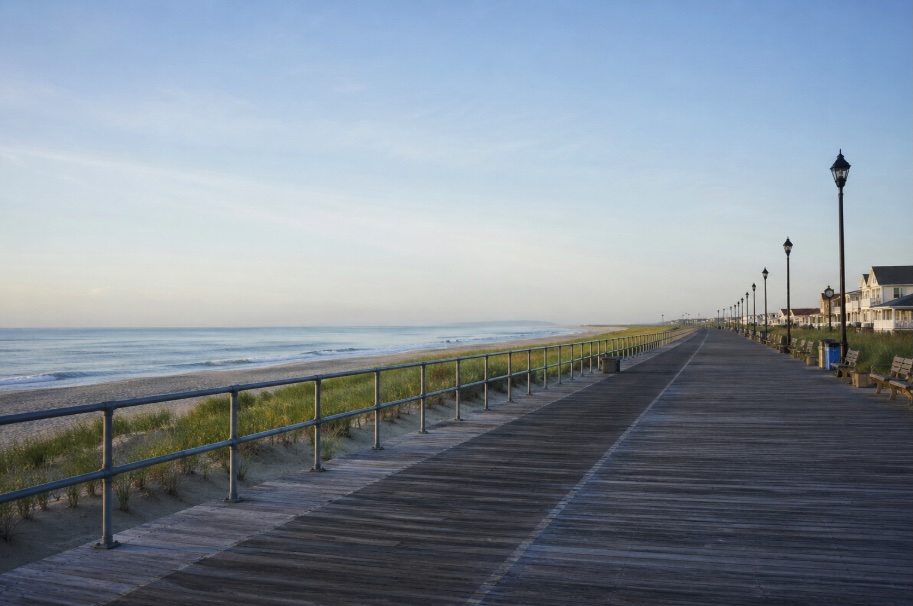 A wide, photorealistic view of the New Jersey Shore on a calm spring morning, featuring an empty wooden boardwalk stretching into the distance alongside a quiet beach. Gentle ocean waves roll in under soft natural light, with muted blue and green tones creating a serene, peaceful atmosphere.