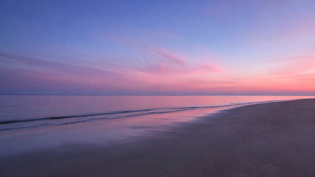 A landscape photo of a wide open beach at dusk, soft lavender and blue sky over calm water, minimal detail, no people, no text, still and reflective mood.